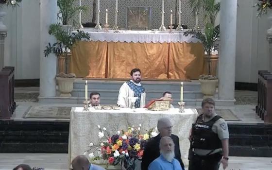 Priest stands at altar while acolytes kneel and police officer scans crowd. 