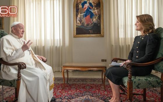 Pope Francis and Norah O'Donnell seated opposite one another, with painting and draped windows in background. 
