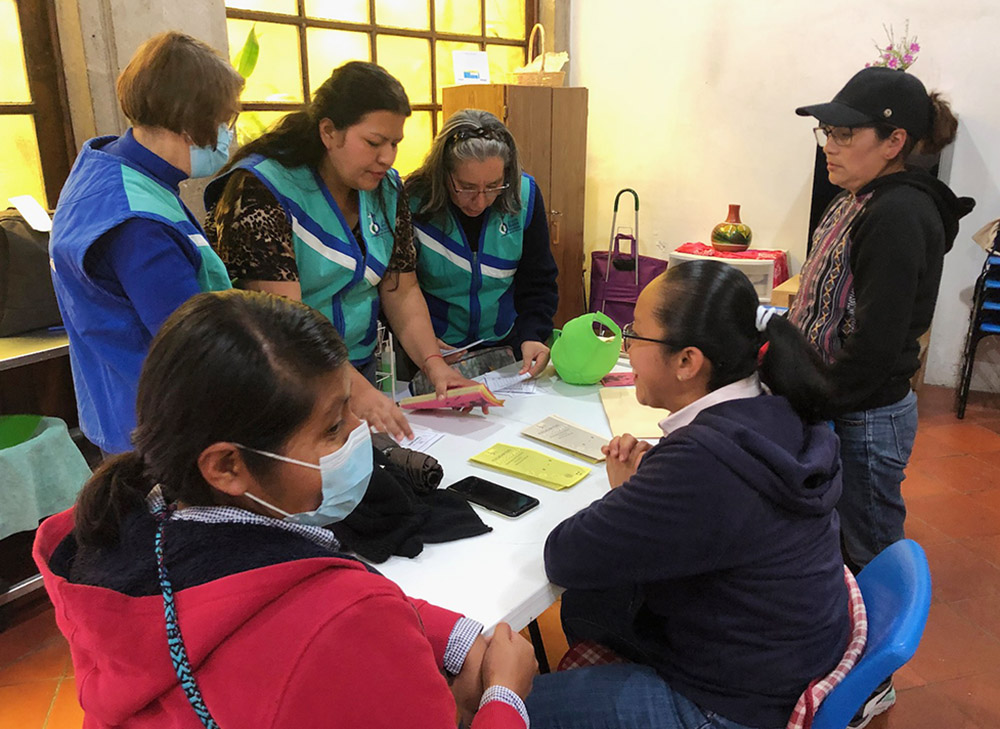 Junto a una mesa de trabajo, las hermanas oblatas Rodríguez, Martínez y Paz, con chalecos azules, se preparan para la sesión diaria de acercamiento en las calles, mientras los voluntarios y el personal observan.