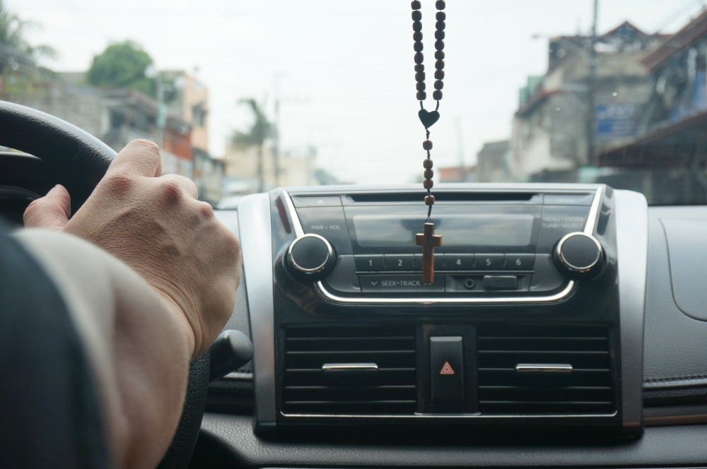 A person driving in a car with a rosary hanging from the rearview mirror (Unsplash/Darla Dela Rosa)