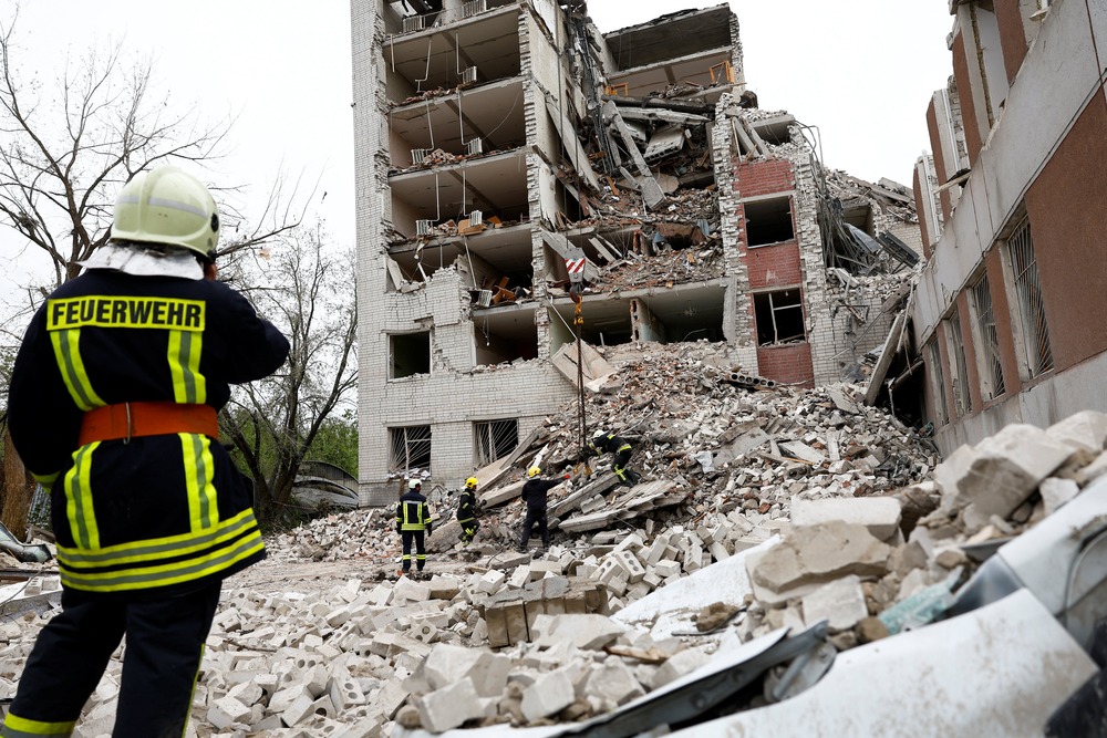 A first-responder, standing on rubble, faces a bombed-out building.