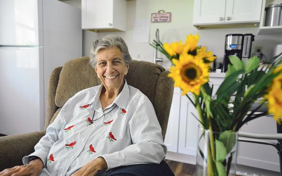 An older white woman sits in a chair smiling at the camera next to a vase of sunflowers