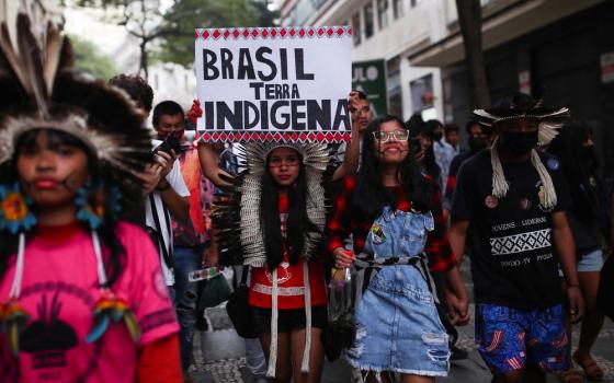 People wearing indigenous garb and ornamentation march in street; one person holds sign reading 'Brazil is Indigenous Land'.