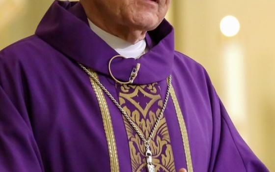 Archbishop, vested in purple, folds hands while preaching. 
