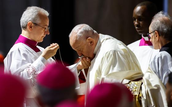 Pope Francis pictured in profile kissing pectoral cross held by aide. 
