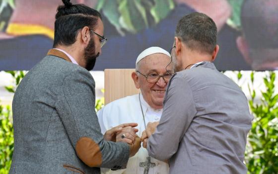 Pope Francis, seated, smiles and takes the hands of the two men. 