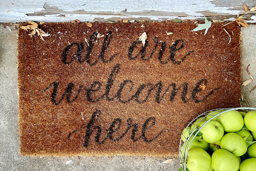 A welcome mat that says "All are welcome here" sits next to a basket of green apples (Unsplash/Leeann Cline)