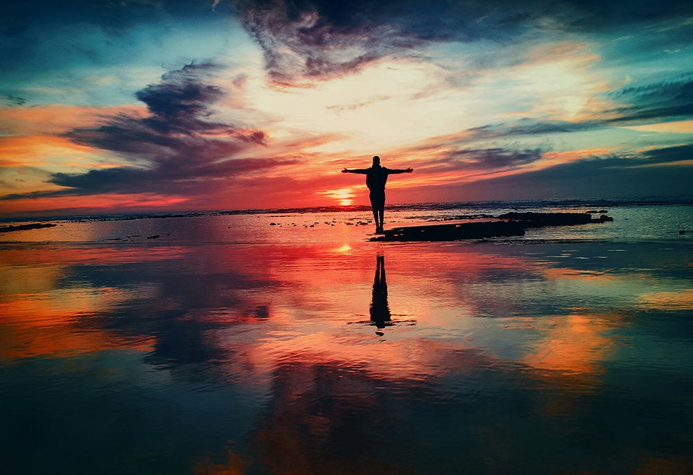 A person stands against a colorful horizon, surrounded by water. (Unsplash/Mohamed Nohassi)