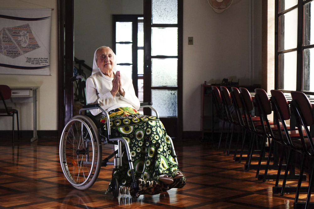 The sister sits in wheelchair holding up prayer hands. 