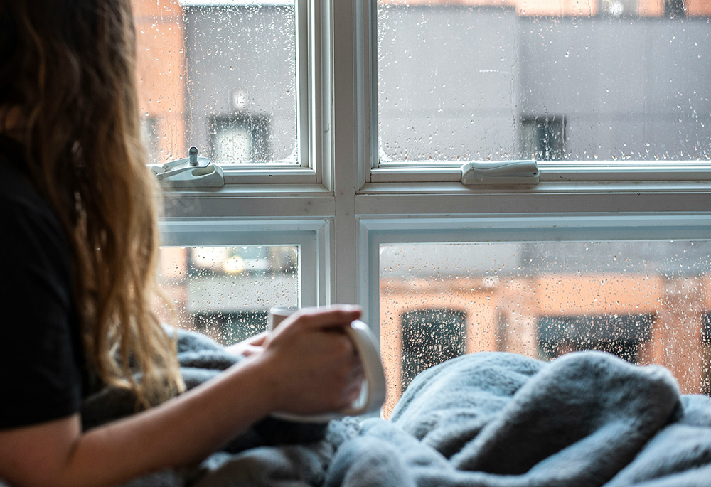 A woman holds a mug while looking out a window during daytime rain outside. (Unsplash/Amin Hasani)