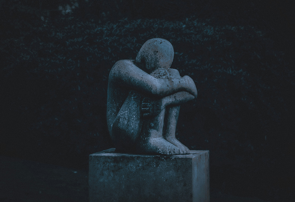 A sculpture depicts a crouched figure holding themselves, their head burrowed against their knees, arms crossed around their legs, in a dark outside setting (Unsplash/K. Mitch Hodge)