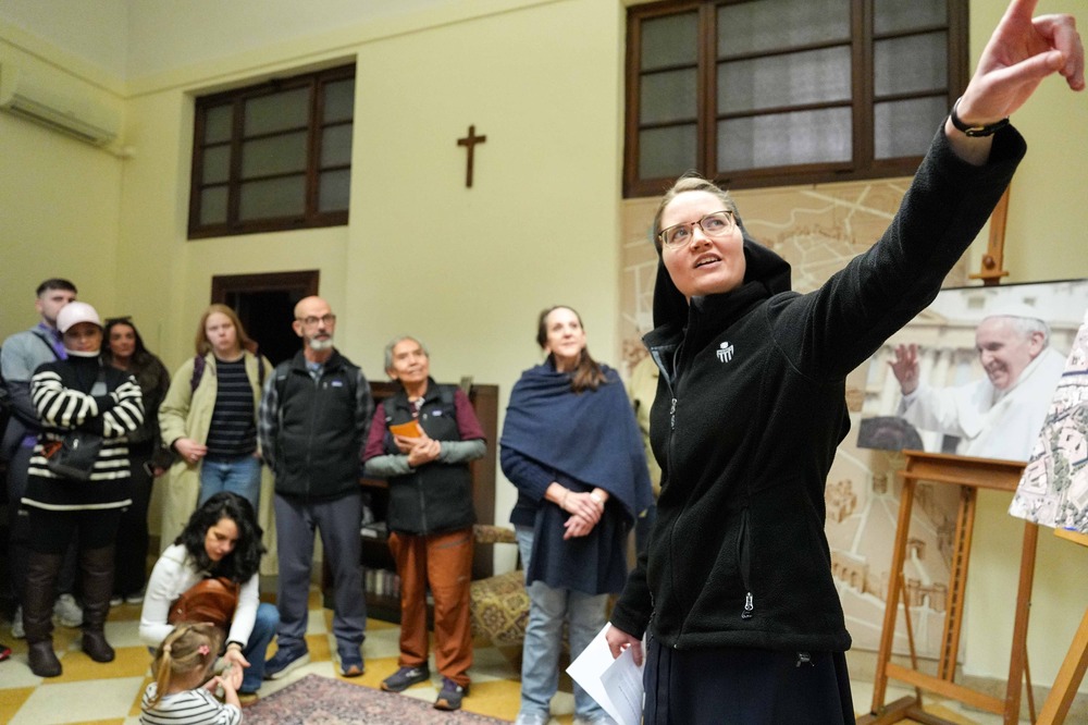 The sister foregrounded pointing out of frame, in background is small group assembled in classroom-like setting.