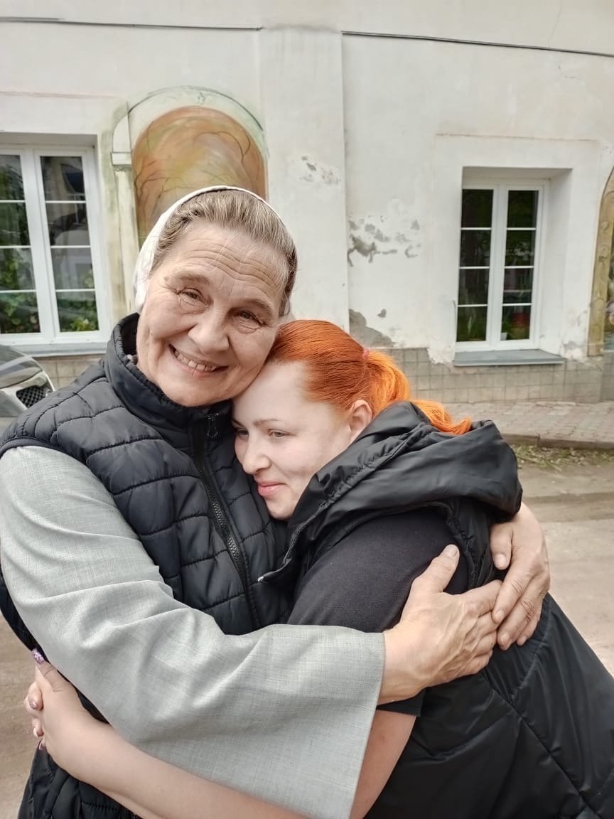 The sister smiles at camera embracing woman who leans into her arms.