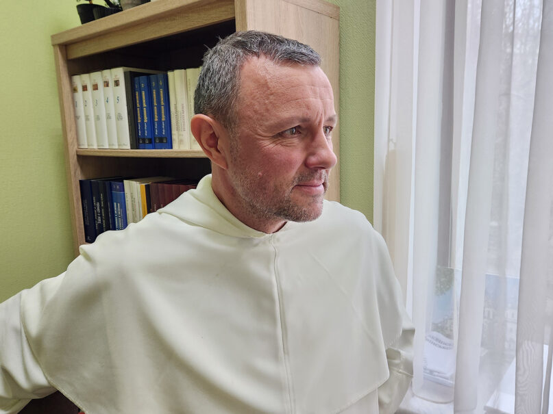 The priest stands wearing white habit staring out of window. 