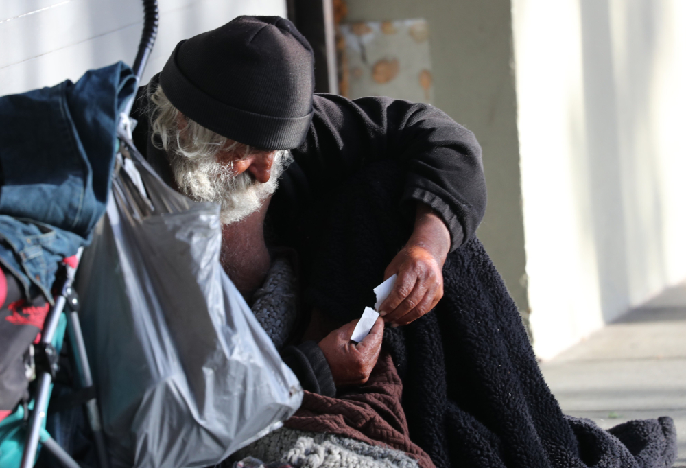 A homeless man holding two pieces of paper in his hands sits with his belongings along a street in San Francisco May 19, 2024.