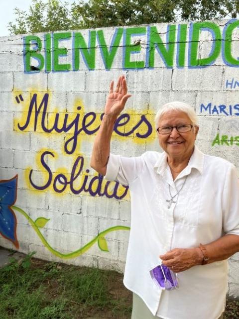 Sr. Virginia Bahena stands in front of a mural and waves.