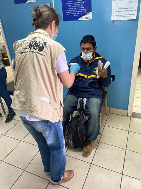 Sister Dondé, standing in a waiting room, offers a drink to a migrant man who is sitting.  