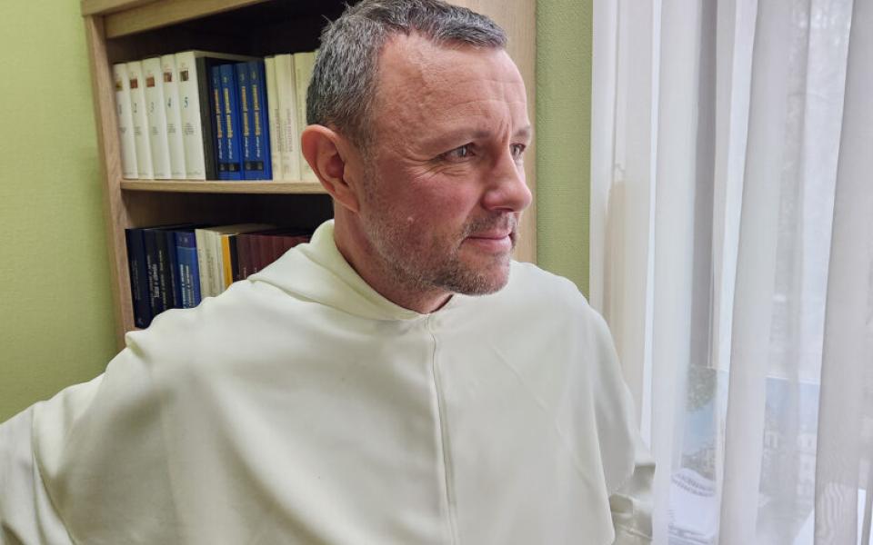 The priest stands wearing white habit staring out of window. 