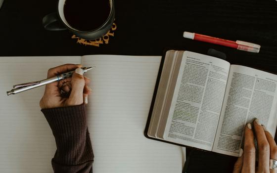 An photo illustration displays an overhead shot of a woman writing in a notebook, referencing a Bible. (Unsplash/Kelly Sikkema)