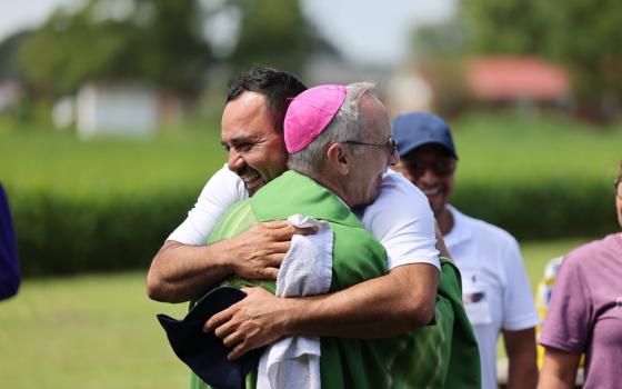 The bishop, vested in green, hugs a smiling man; in the background is a field of tall green stalks, possible corn. 