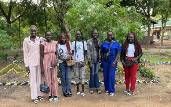 Seven young women stand in  row. In the background are trees, garden beds, and buildings.