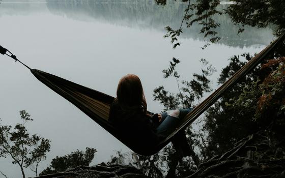 A woman sits on a hammock facing towards a lake in a forest setting (Unsplash/Priscilla Du Preez)