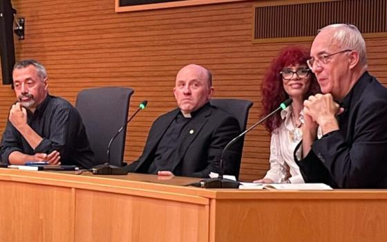 The four sit at a wooden desk with microphones. 