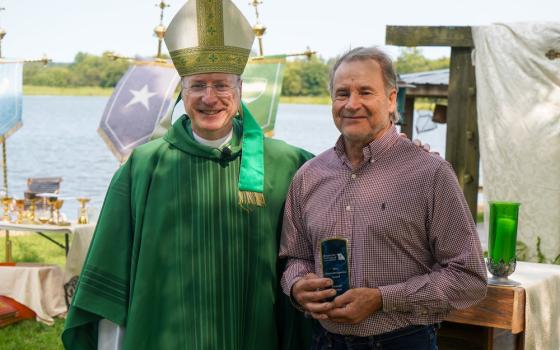 The bishop, vested for ordinary time and wearing mitre, stands with his arm around Kremer who holds his award plaque.