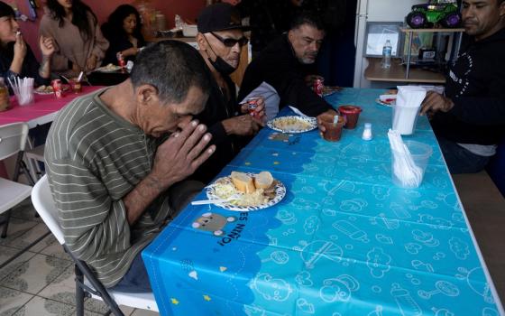 Group of people sit at long tables; foregrounded man bends his head in prayer. 