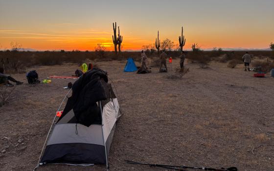 Desert panorama lit with orange hues of sunset, foregrounded is a tent and people sitting and walking.