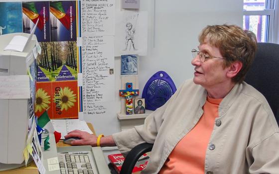 An undated photo of Patty McCarty at her NCR newsroom desk. A partial list of saints is on the wall and other icons surround her. (Courtesy of Tom Fox)