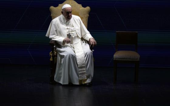 Francis sits on chair, highlighted by light, surrounded by shadow.