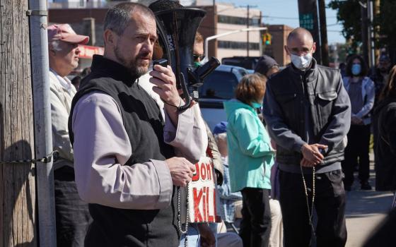 The priest stands holding megaphone and rosary.