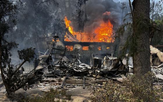 Fram filled by smoke and bordered by trees; centered is blackened shell of two-story house still burning.