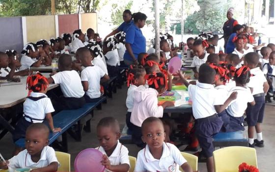 Large group of children sit along tables in covered outdoor area.