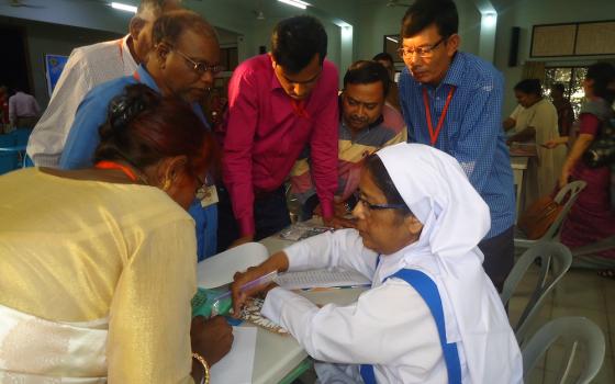 A habited sister in white and blue points to papers on a desk while others gather around to listen