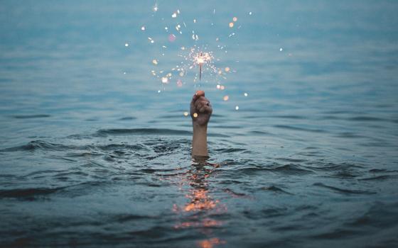 Image of a person holding a lit sparkler above the water (Unsplash/Kristopher Roller)